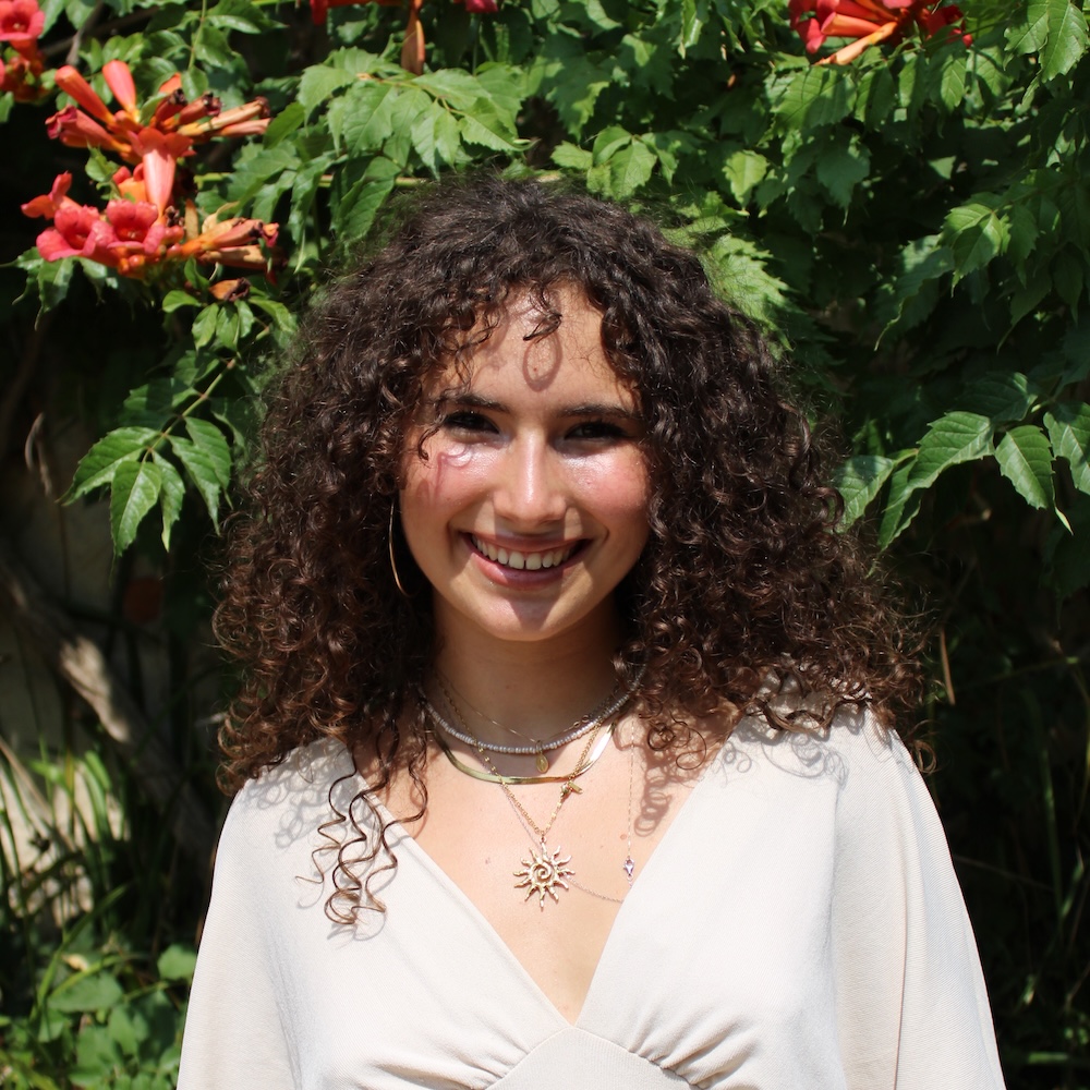 Person with long curly brown hair, wearing a cream dress smiling at the camera.