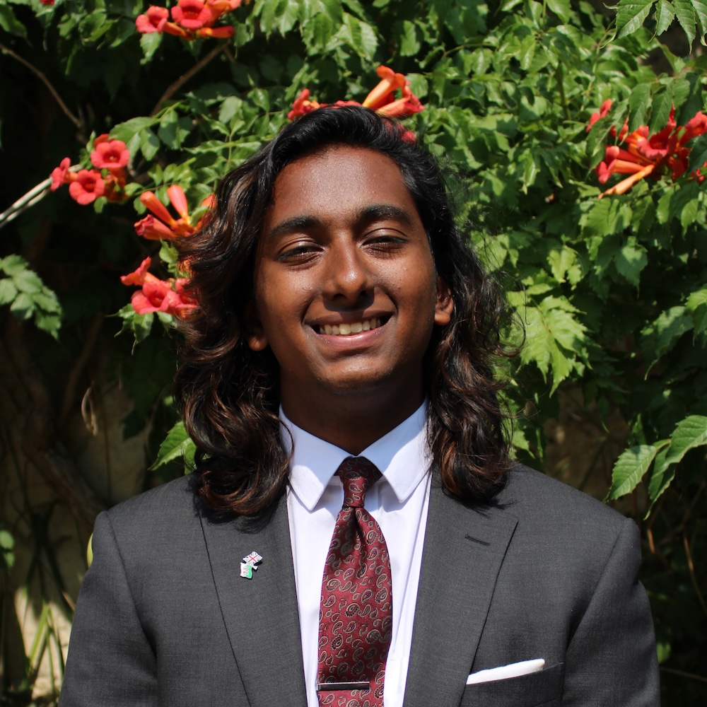 Person with shoulder length wavy brown hair, wearing a grey suit, white shirt and red tie, smiling at the camera.