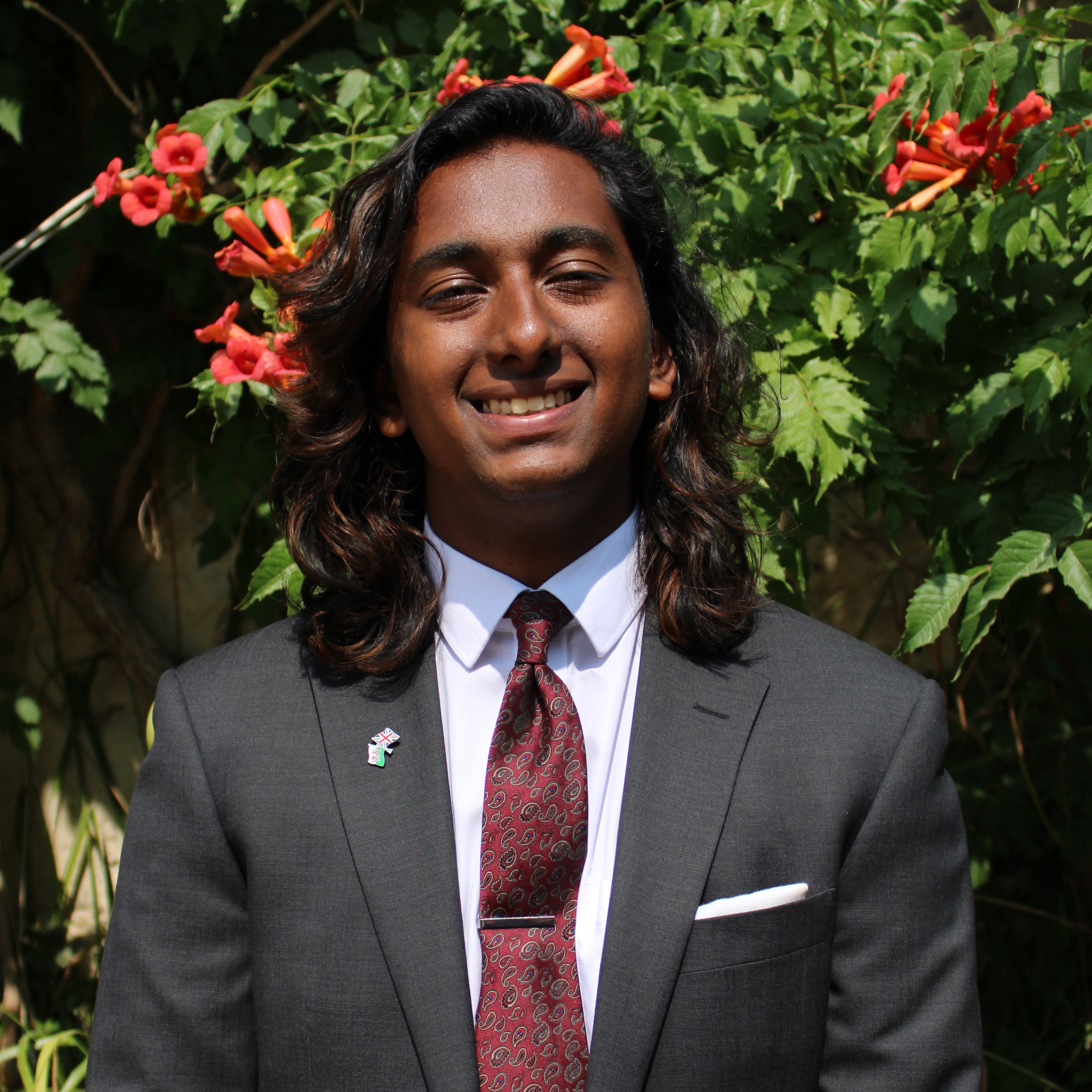 Person with shoulder length wavy brown hair, wearing a grey suit, white shirt and red tie, smiling at the camera.