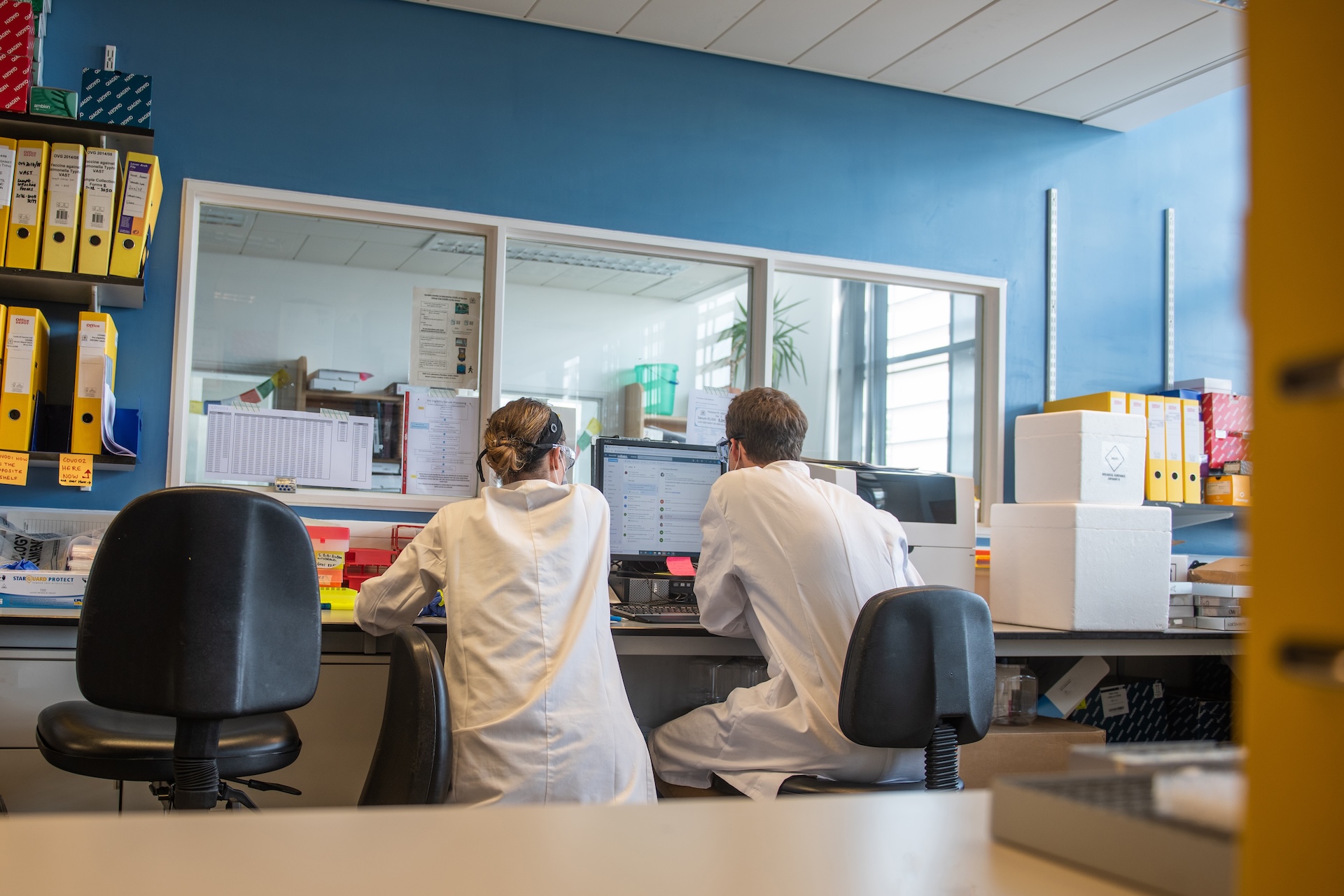 Two people sat in a lab wearing white lab coats, looking at research material