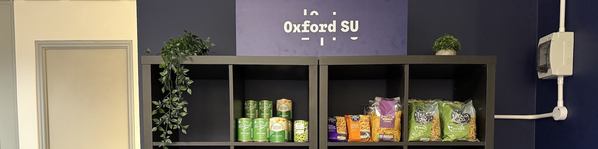 Shelves with food items against a navy blue wall, with a sign on top featuring the Oxford SU logo in white