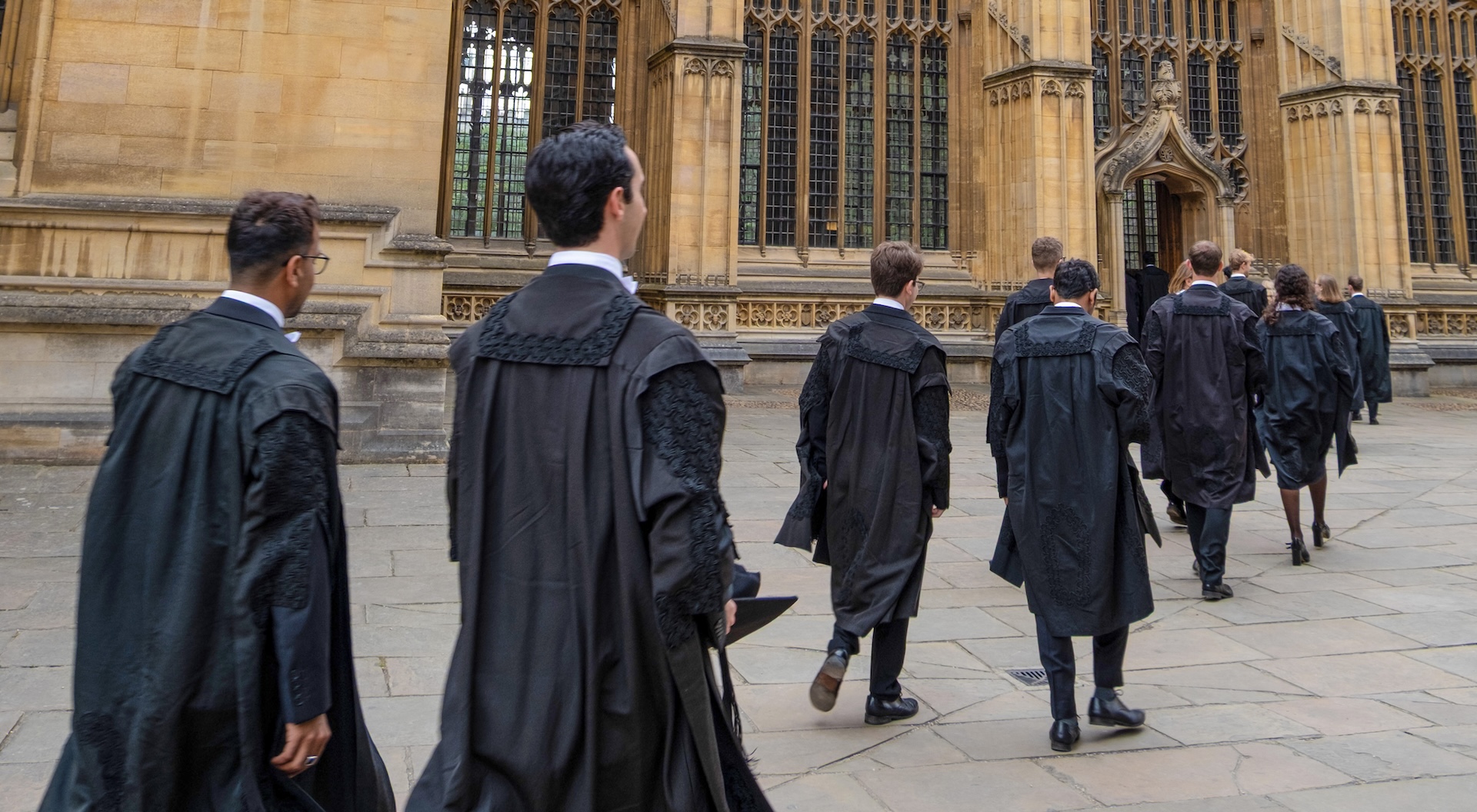 Group of students in black formal attire walking in pairs towards a historic stone building in Oxford.
