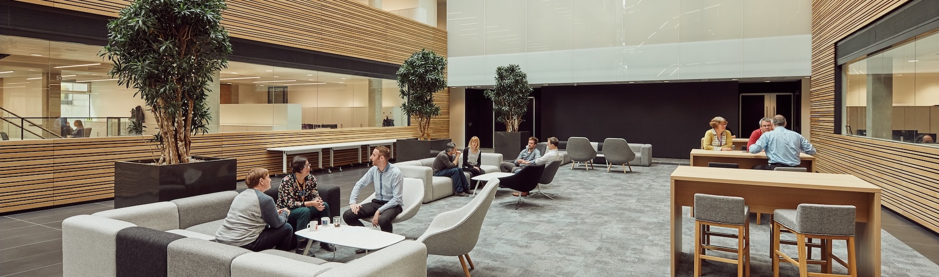 Group of people sat around tables talking in a high ceiling university lobby area.