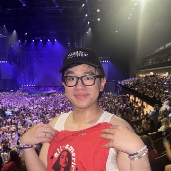 Person with thick black glasses and short black hair standing in a stadium audience packed full of people