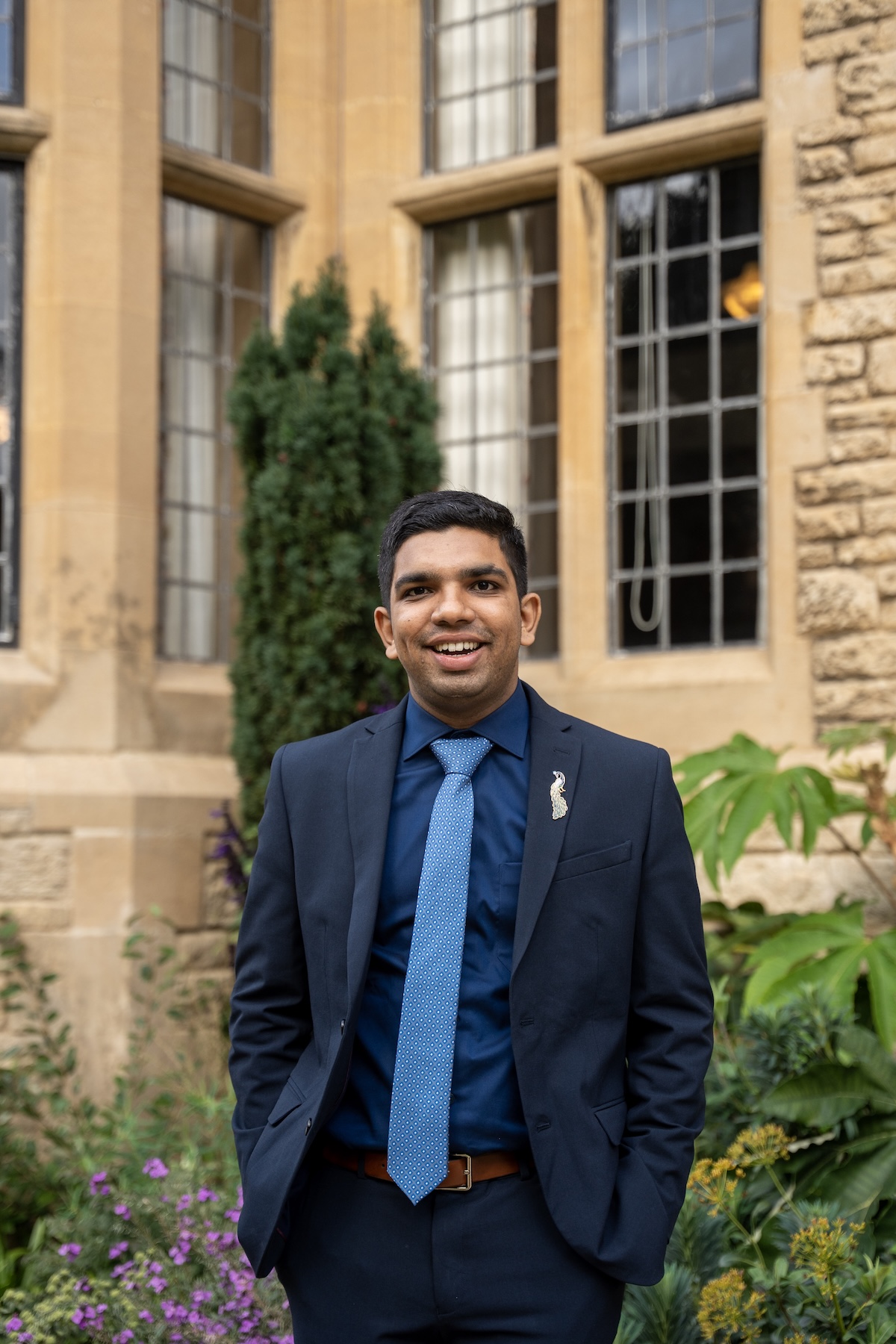 Portrait of person wearing smart dark suit and shirt, with a blue tie standing in front of an old university stone brick building