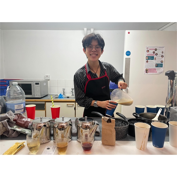 person with short dark hair and glasses, wearing a stripey black shirt serving coffee behind a cafe counter