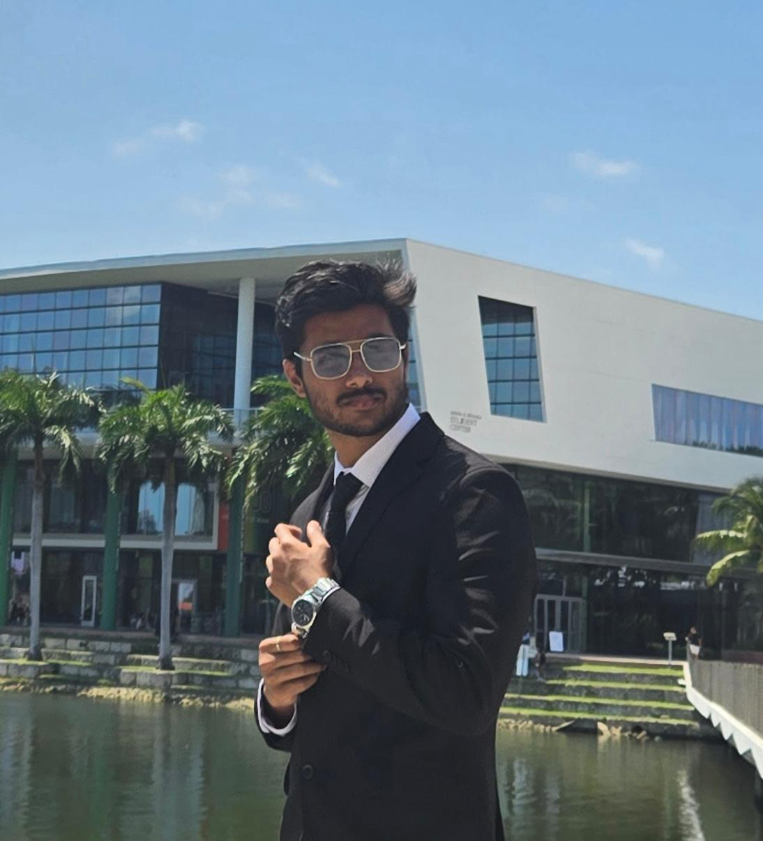 Portrait of person with black wavy hair, a beard wearing silver sunglasses and a black suit. They are adjusting their silver watch on their wrist.