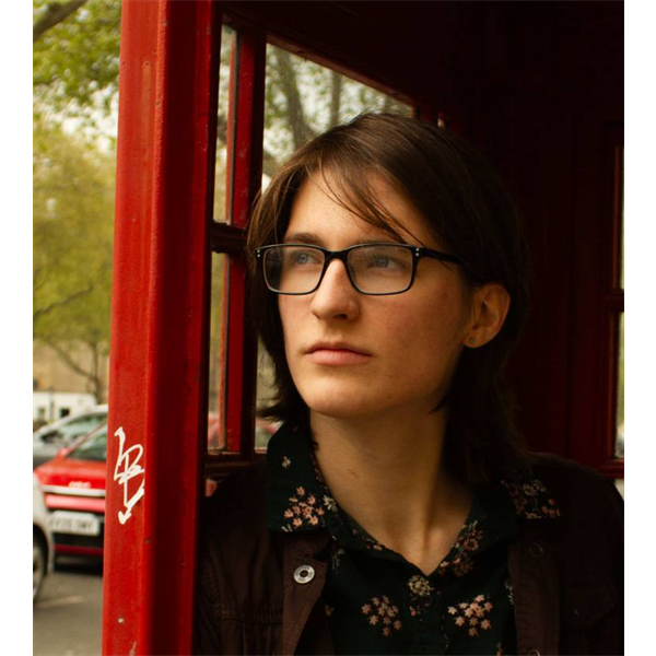Portrait of person with long dark brown hear, wearing black rectangular glasses, standing in a red british telephone box