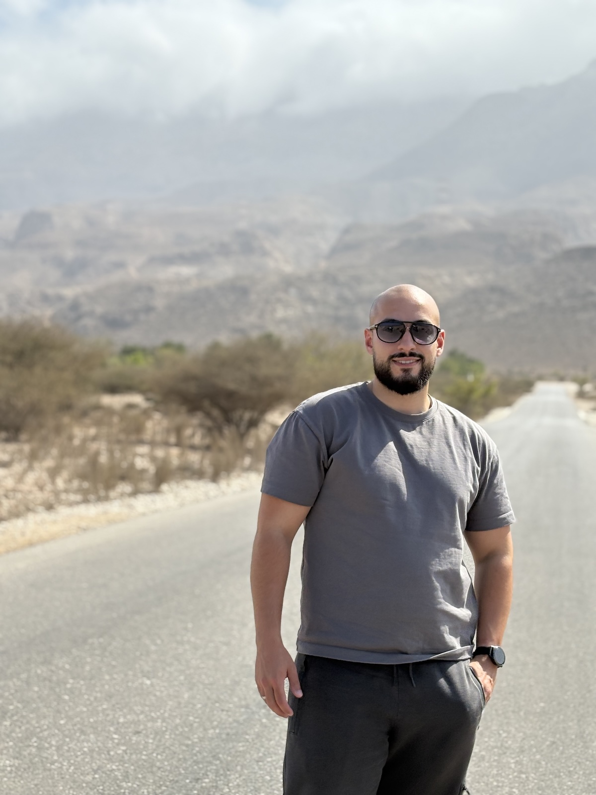 Portrait of a bald man with a beard, he's standing on a road in nature, wearing sunglasses and a grey tshirt